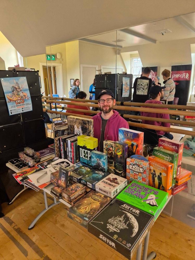 Ancient Robot Games board game shop staff member smiling behind a stall full of board games and RPG zines.