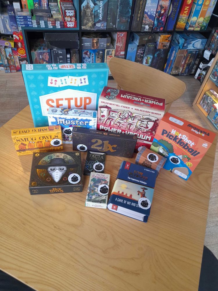 Several games on a table in the Edinburgh board game shop Ancient Robot Games. The games include Setup, Muster, Power Vacuum, Hotshot Pickleball, Smug Owls, 21X, Hex effects, Tacta, Don Quixote The Ingenious Hidalgo, For the Emperor and Game Chest. 