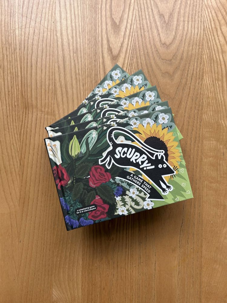 Copies of the TTRPG book Scurry on a table at the Edinburgh board game shop Ancient Robot Games. The cover has illustrations of a mouse and flowers.