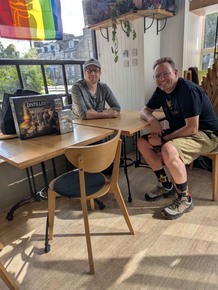 The owner of the board game shop Ancient Robot Games and the game designer Dave Beck sitting by a table and smiling. There is a copy of the board game Distilled on the table.