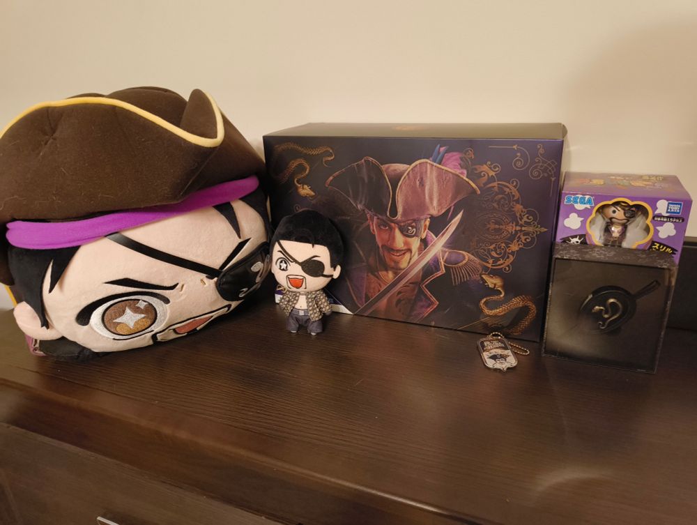 A photography of various Majima Goro merch: a jumbo plushe, Tiny plushie, collectors edition box, eyepatch, keychain and pirate barrel toy. All of it on dark brown cabinet