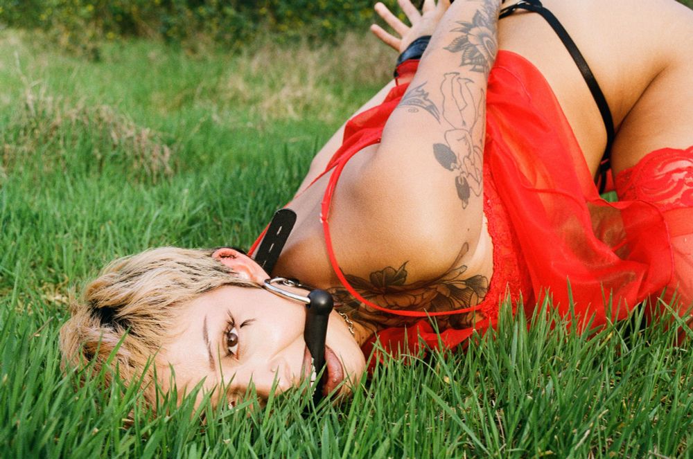 Bebecita is laying face down ass up, on the grass as their mouth is propped open by a bit gag and their arms restrained behind their like back.