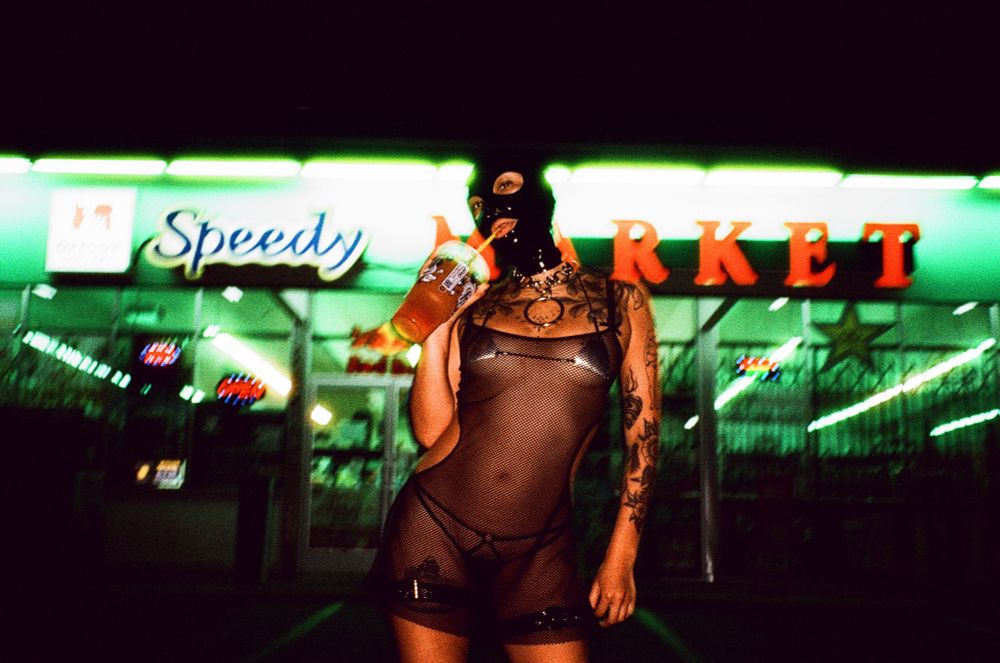 Bebecita stands in front of a fluorescently lit corner store at night. They stare at the camera while they drink from a large transparent soda cup. They’re adorned in a black latex hood and a mesh mini dress.