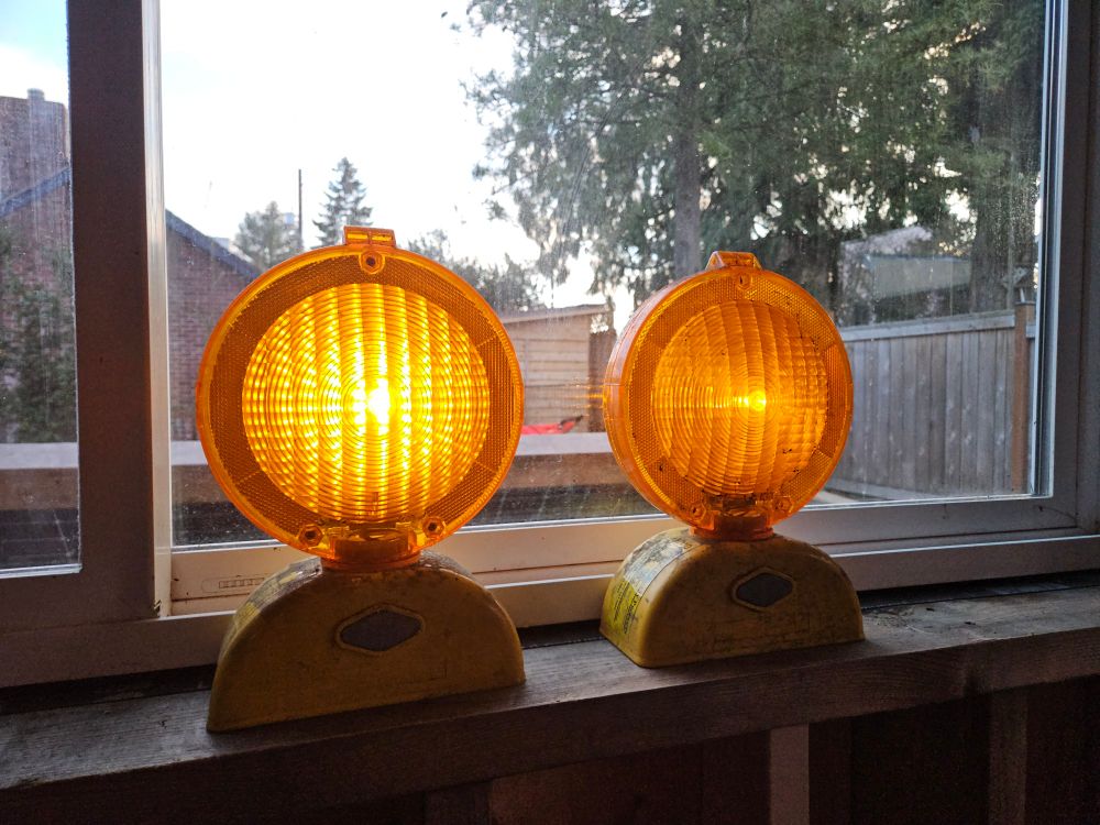 Two amber barricade warning lights with round, textured lenses glow on a wooden windowsill. Their yellow bases show signs of wear. A window behind them reveals a dimly lit backyard with a fence, trees, and a shed.