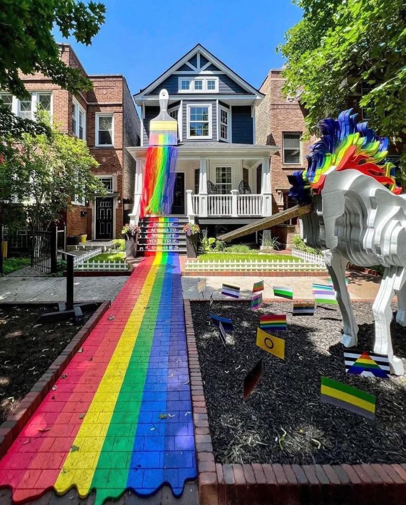 photo of a house in Boystown, a neighborhood in Chicago. the house has a giant paint brush with rainbow paint, running down the steps of the home and across a brick path. The steps says "Don't be afraid to show off your true colors." in the foreground is a unicorn with a rainbow mane. the yard is populated with various versions of Pride flags.