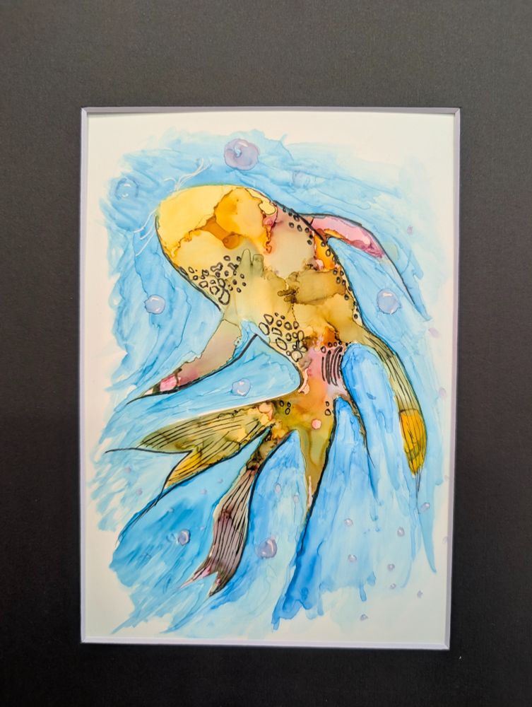 his artwork depicts a colorful fish rendered in a fluid, abstract watercolor style. The fish is shown in motion, curved slightly as if swimming upward through water. Its body is painted in a mix of warm, earthy hues—yellows, golds, oranges, and browns—blended with soft washes that bleed into each other, creating an organic, marbled effect. Darker speckles and spots decorate the top half of its body, giving the suggestion of scales or natural patterning.

The fish’s fins and tail are long and flowing, trailing gracefully behind it. They extend outward in multiple directions, with delicate ink outlines adding definition. The fins are painted in tones of pink, yellow, and black, blending into the watery blue background. Some fins are lined with narrow strokes that hint at texture or ribbing.

Surrounding the fish is a loosely painted wash of blue, representing water. The watercolor is light and translucent in areas, giving the background a soft, almost swirling effect. Scattered across the scene are small, round, translucent circles in pale purples and blues, resembling air bubbles rising through water.

The entire scene is bordered by a narrow white frame and set against a dark background, which helps the light colors of the painting stand out. The overall style is loose and expressive, with visible brushstrokes, ink lines, and watery color blending adding a sense of motion and life to the image.