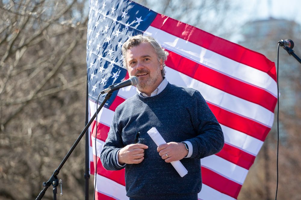 Mass Dems chair speaking at Boston rally in front of large American flag. 