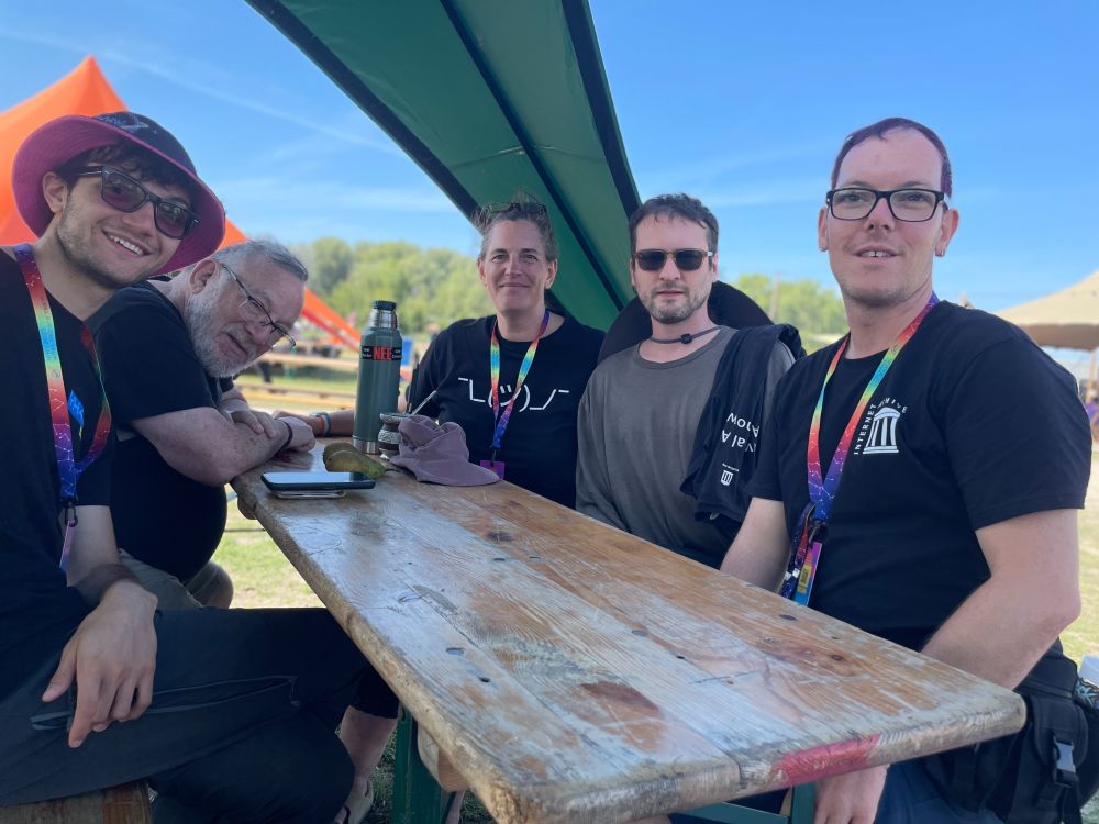 Five people sitting around a wooden table under a green shade tent. Two men on the left and a woman and two men on the right.