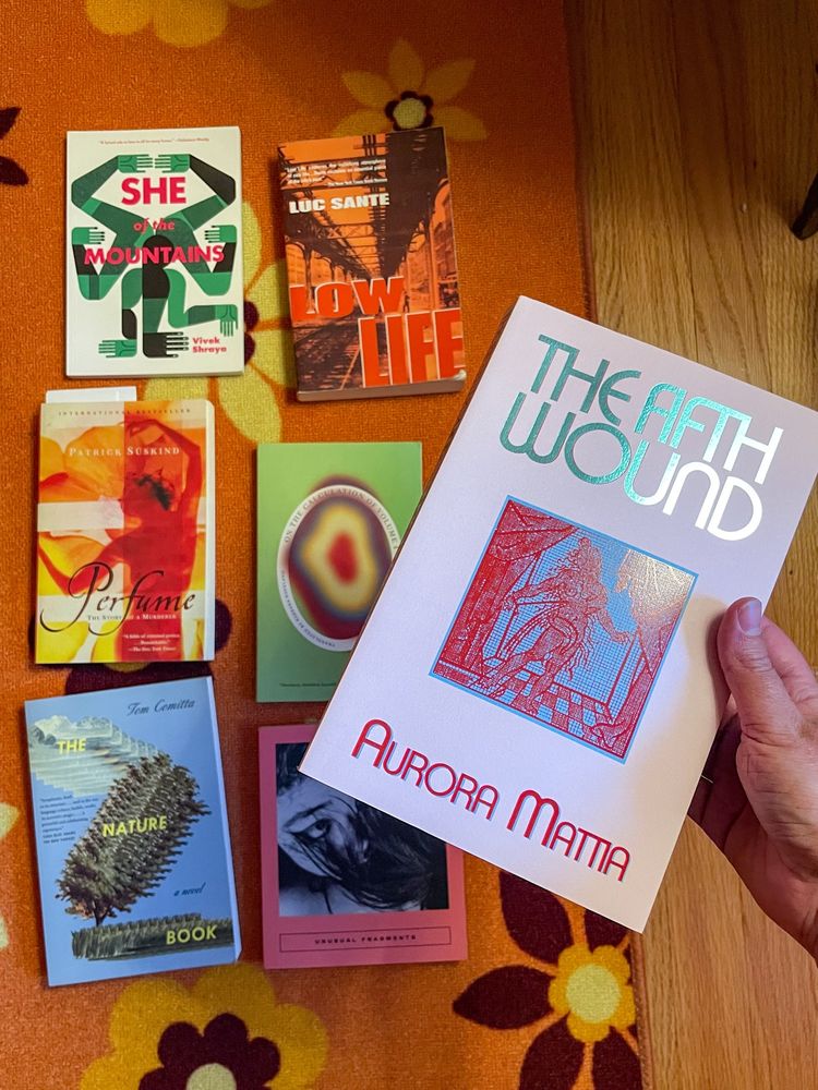 Books laid flat on a floral carpet. She of the Mountains by Vivek Shraya, Low Life by Lucy Sante, Perfume by Patrick Süskind, On the Calculation of Volume 1 by Solvej Balle, The Nature Book by Tom Comitta, Unusual Fragments: Japanese Stories, and The Fifth Wound by Aurora Mattia.