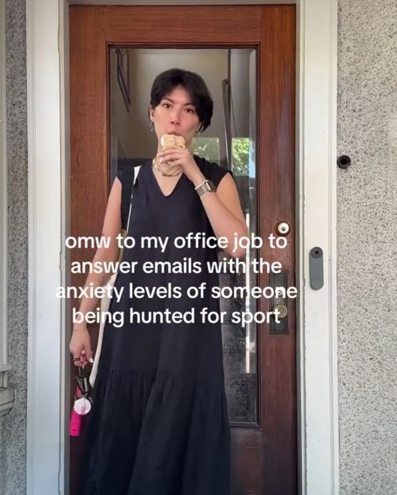 person with dark hair and a long black dress standing outside a door and eating a sandwich (I think it's a sandwich? maybe a burrito? idk) with a caption on top saying "omw to my office job to answer emails with the anxiety levels of someone being hunted for sport" 