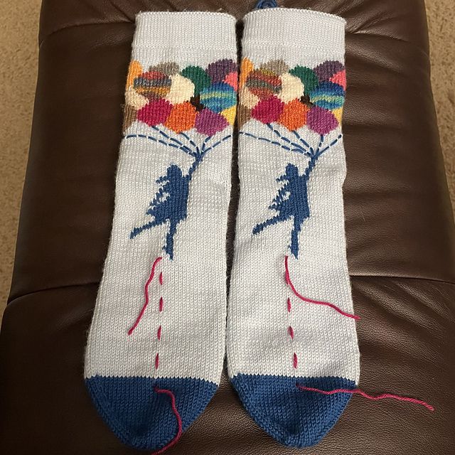 A pair of handknit socks laid flat displaying the front side. They are light blue with darker blue toes. Depicted by using intarsia, is a girl floating away holding a bundle of multi-colored balloons. There is a strand of fuchsia yarn temporarily placed vertically as a row counter, and has since been removed. The pattern was the qualifying round for Sock Madness 17 in 2023. It is called Leicht by Annette Gürtner.