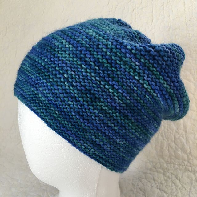A handknit garter stitch slouchy hat in variegated blue & teal. Displayed on a white styrofoam head form. Background is white quilted fabric. Pattern is Rikke Hat by Sarah Young.