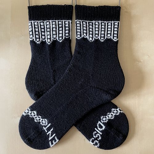 A pair of handknit socks in black and white, light colored faux wood background. The colorwork at the cuff is a representation of the collar worn by Supreme Court Justice Ruth Bader Ginsberg.