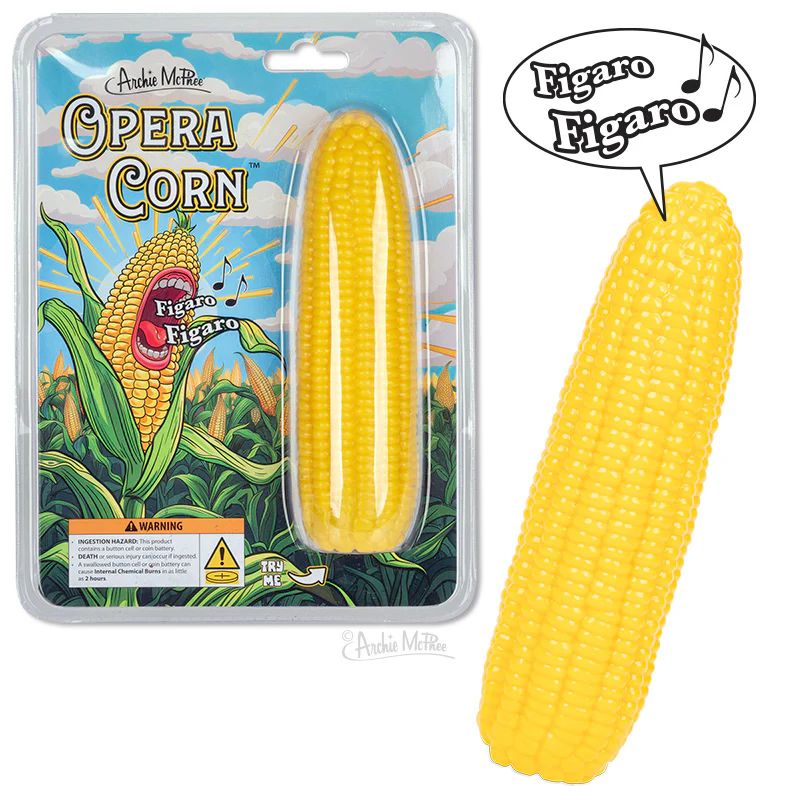 "Opera Corn," designed by Archie McPhee - a bright yellow corn cob that, when activated, plays the famous aria "Figaro" from The Barber of Seville along with other opera related sounds. The corn cob is displayed in its packaging, which is illustrated with vibrant imagery of corn plants against a blue sky, featuring a cartoonish corn cob singing the word "Figaro."
