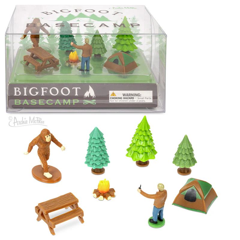 A toy set titled "Bigfoot Basecamp" featuring miniature figures and accessories, including Bigfoot, a camper, a campfire, a tent, picnic table, and several trees. The items are displayed in a transparent box.