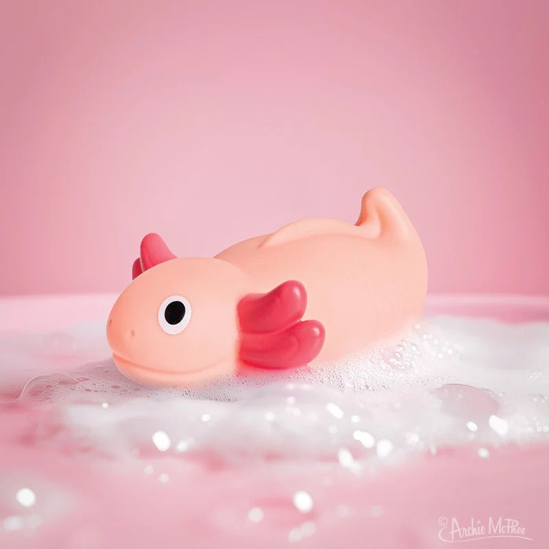 A light pink rubber axolotl toy floats in soapy water, surrounded by bubbles against a soft pink background.