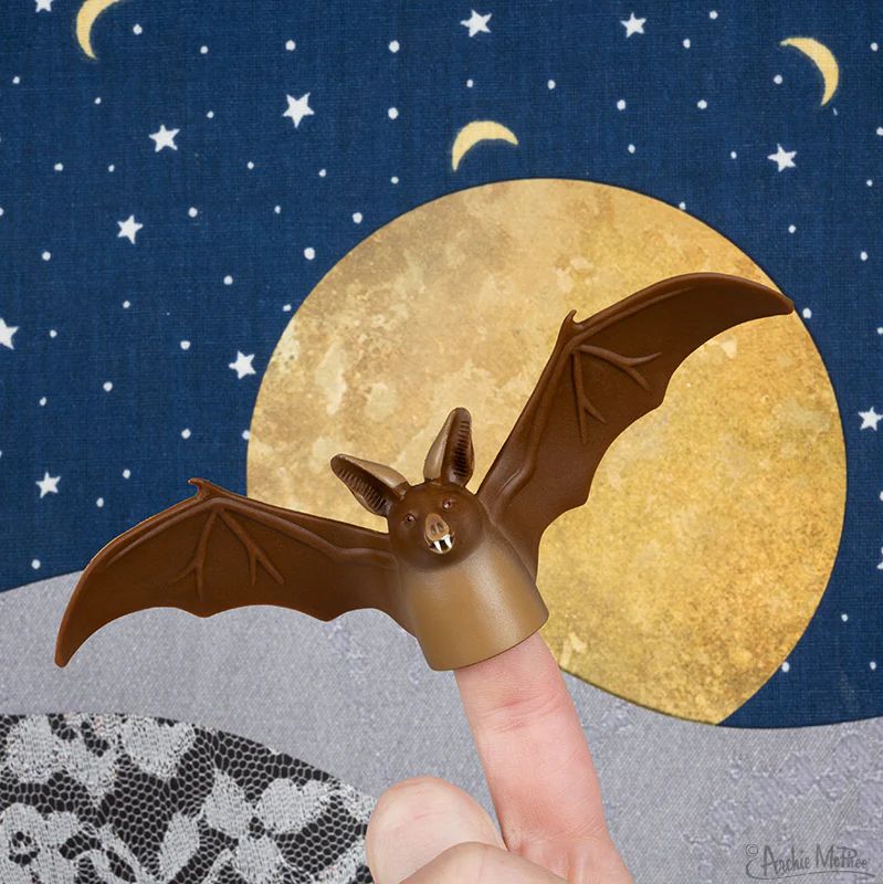 A finger puppet shaped like a bat is shown, placed on a person's finger against a backdrop featuring a large moon and a starry night sky decorated with crescent moons. 
