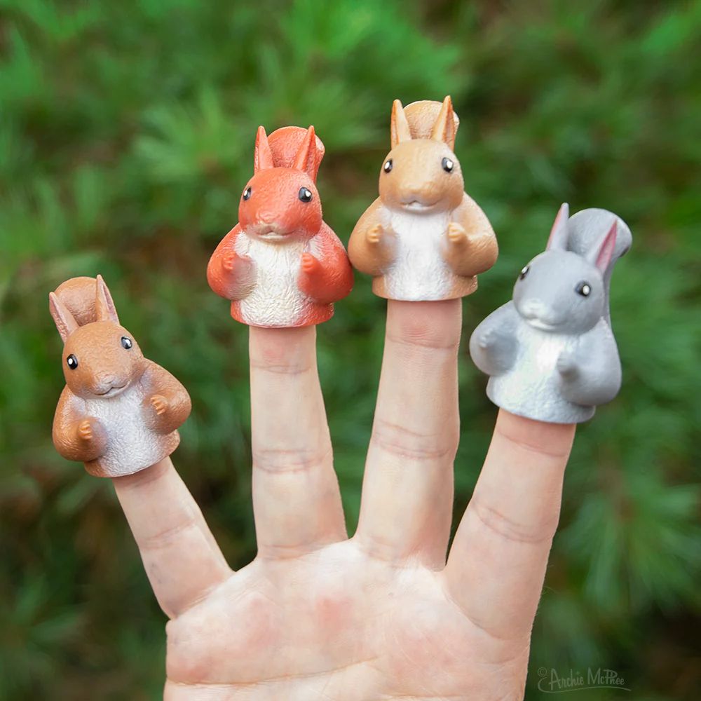 A hand with four finger puppets designed to resemble squirrels, featuring different colors: orange, brown, and gray. The background consists of greenery.