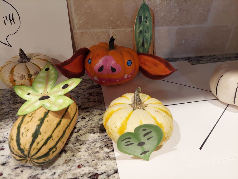 Several small decorative pumpkins and gourds of varying shades from white to orange, some light orange with green stripes. The orange pumpkin has a pink, pig-like nose, blue eyes, tusks, and floppy ears. It's meant to lookike a bokoblin from The Legend of Zelda Breath of the Wild. The rest have leaf shaped green masks with faces. These are meant to look like the Korok spirits from the same game.