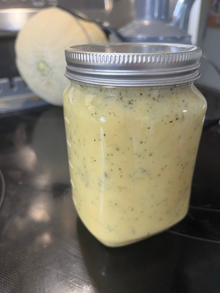 A plastic jar with salad dressing, which is a light yellow with green flecks of herbs 