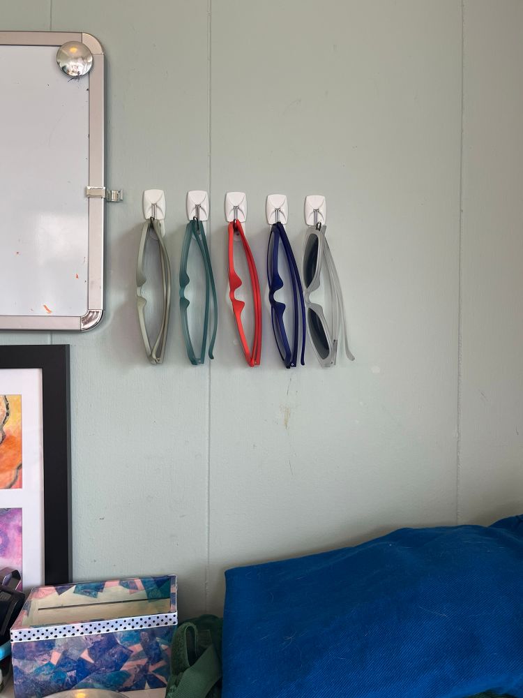 Five pairs of sunglasses hanging in a sea foam green wall on small hooks, above a shelf with other stuff on it. The sunglasses are, left to right opaque mint green, sea glass green, opaque orange, royal blue, and frosted white 