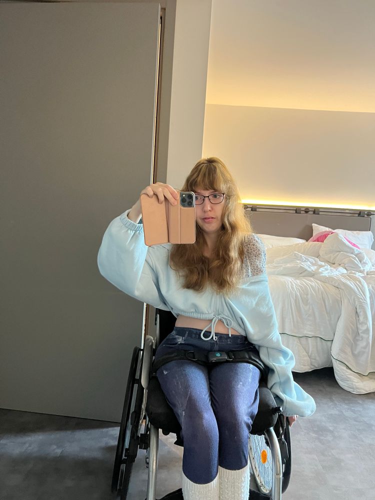 Mirror selfie of femme woman in wheelchair, long brown/blond hair, glasses, baby blue crop top hoodie, blue pants, white leg warmers 