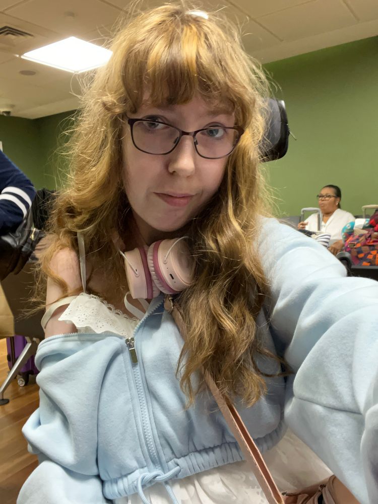 Selfie of a femme woman in wheelchair, white top, bang blue hoodie. Brien/blond long hair 