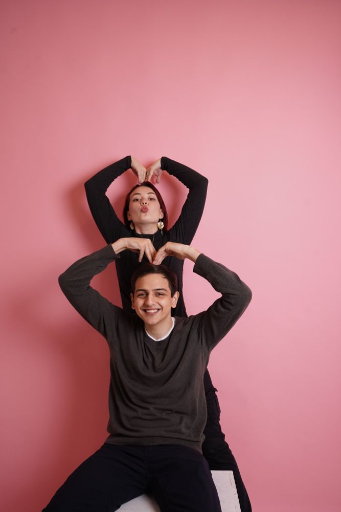 Woman with black turtleneck lifts her arms up above her head to make a heart shaped and a kissy face. A man in front of her is doing the same pose but smiling.