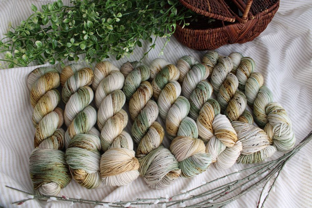 8 skeins of yarn in a row. There is a picnic basket with greenery flowing out of it above the yarn and some pussy willow branches below the yarn. Each skein of yarn is dyed in a variegated fashion with shades of white, leafy green, golden straw, and speckles of brown. Each skein looks a little different as each has a different fiber content. 
