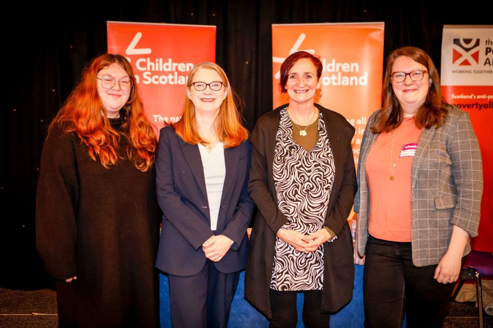 Four people stand together, smiling at the camera. Behind them are two red pop-up banners that read 'Children in Scotland'.