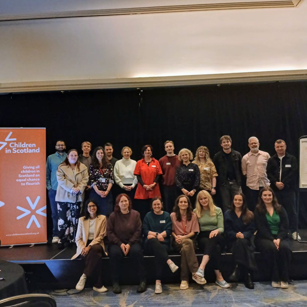 A group of Children in Scotland staff pose for a group photo on the stage of a conference room.