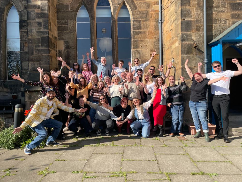 A group of around thirty people stand outside of an old church smiling and waving their hands in the air