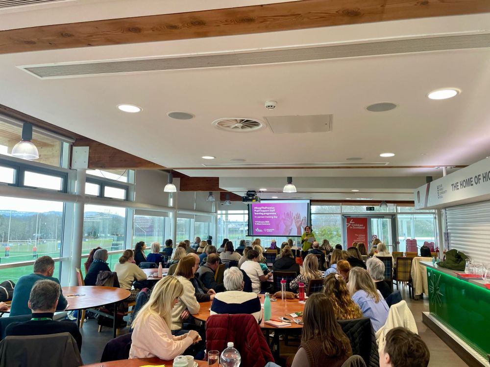 A room full of seated people facing a presentation screen with a purple graphic pictured. Through the window of the room can be seen a rugby playing field. 