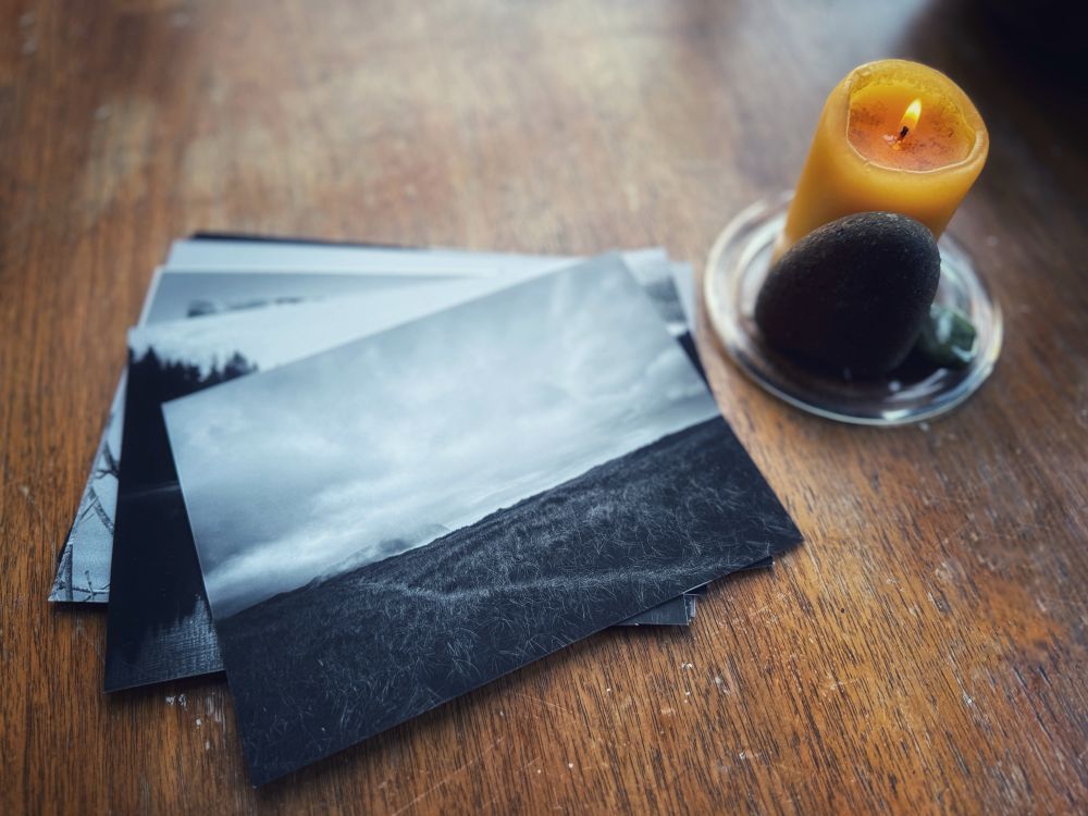 A lit candle next to a beach rock and a shiny green stone. A pile of black and white photos, the top one shows a path through beach grass and a cloudy sky. 