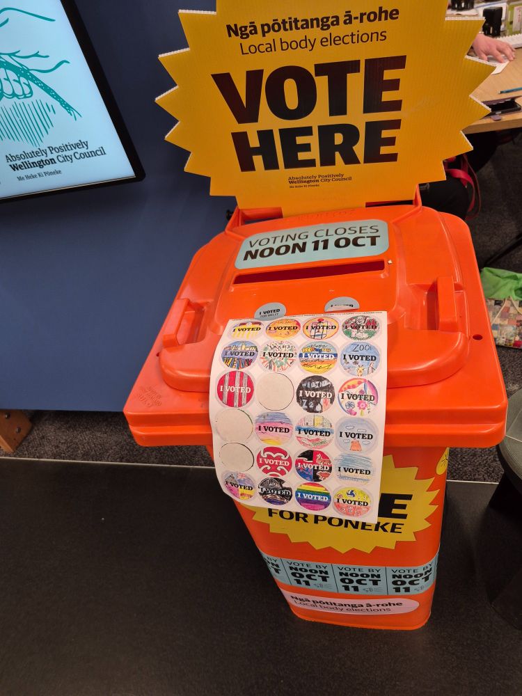Image of orange recycling bin, being used as a voting box. Attached to it is a part used sheet of hand drawn I Voted stickers.