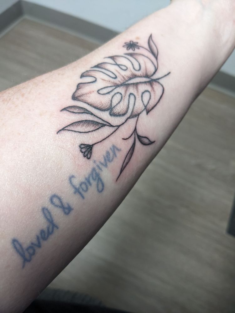 Showing my tattoo on the left arm. My old tattoo says "loved and forgiven" in blue ink. The new one is a monstera leaf, now shaded in, with some extra leaves & flowers added.