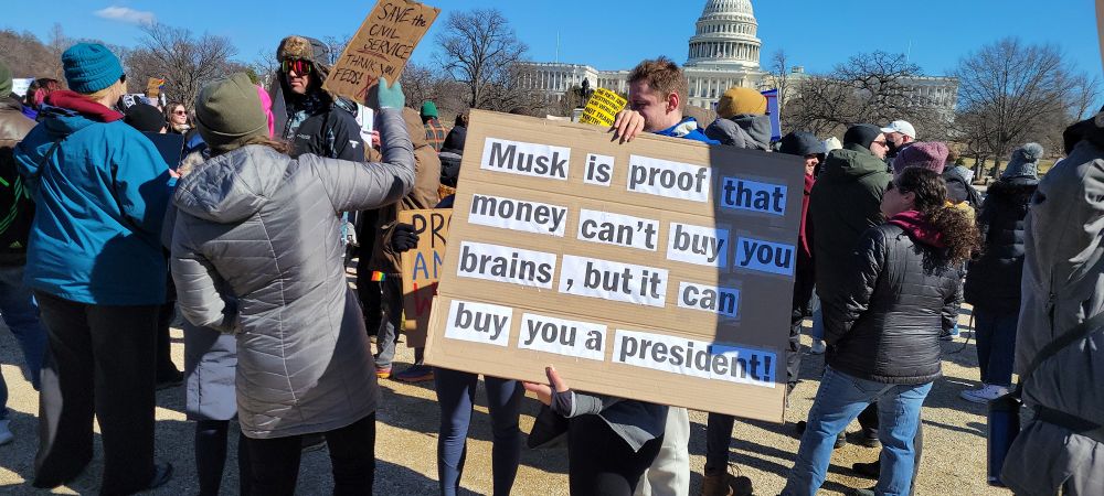 Sign saying "Musk is proof that money can't buy you brains, but it can buy you a president." 