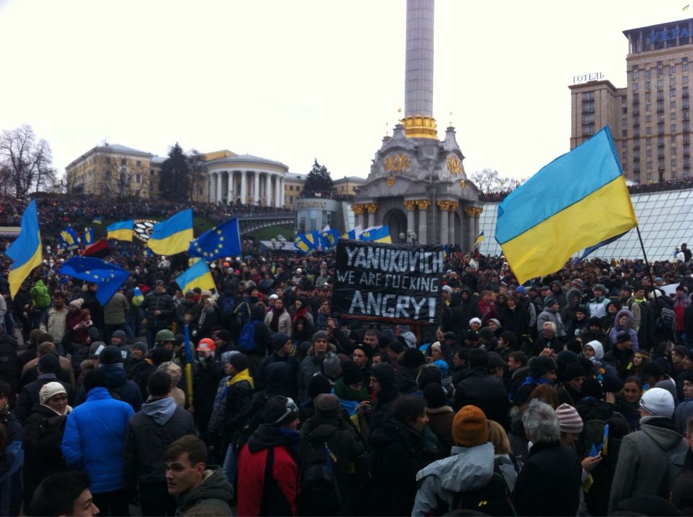 A Euromaidan protest, Ukraine and EU flags waved about with one barely visible OUN flag in the distance. A sign reading "YANUKOVICH WE ARE FUCKING ANGRY!"
