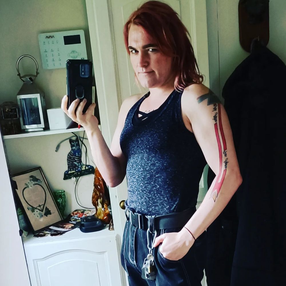 Me by the front door rocking a proper butchy look. Muscle top for to show off, well, the muscles. Ngl, my arms look DAMN good in here. Face in the light, giving a mischeivous look, hand in my pocket, thumb resting on my carabiner