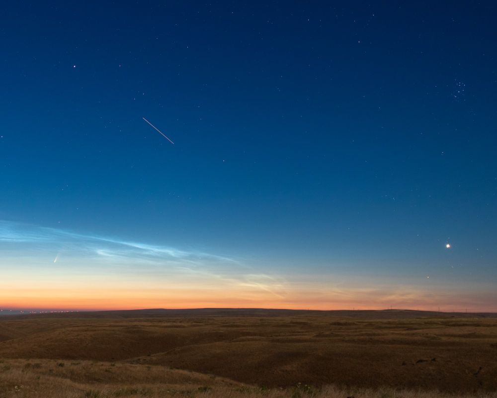 A predawn photo of the sky, but that's not all!
Across the top of the image are many small stars set against a deep blue sky which gradually turns to a gradient of pale yellows and orange of sunrise colors at the horizon toward the bottom of the image. On the far right, and down from the top of the image is the small bright constellation of The Pleiades, and much lower down toward the brightening horizon is the brilliant planet Venus.
From the center of the image, and just up to the left is a bright streak of light, slanted down from left to right. That is the trail of the International Space Station over a few seconds of exposure, as it quickly crosses the sky.
Below that, closer to the changing colors along the horizon is a ghostly baby blue, twisting cloud that stretches from the left of the image to about the center along the horizon. These are rare clouds very, very high in the atmosphere called "Noctilucent Clouds". (I will post a link to a wiki article on them in the comments)
Almost hidden in the advancing dawn colors and the Noctilucent Clouds is a dash of brightness steeply tilted from right, to left. That is the Comet Neowise (Also an article in the comments).
Below the horizon are low, getnly rolling hills as far as the eye can see, in a dull gold color of grasses, lit by the light of the Moon (the moon is not in the image frame). Also, along the horizon, and the the far left, is a large grouping of small red lights in the distance. Those are a huge number of safety lights atop electrical wind turbines.