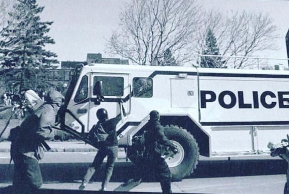 A masked anarchist in a helmet smashing the window of a police vehicle (marked "POLICE") with a water cannon mounted on it.