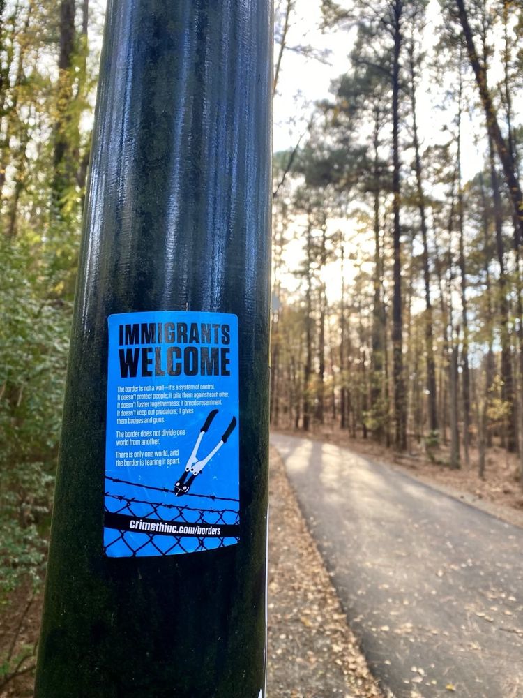 A sticker reading "Immigrants welcome" with an image of boltcutters cutting barbed wire on a pole by a road in the forest.