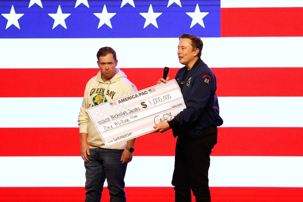 Brandishing an oversize check for a million dollars from "America PAC," Elon Musk poses with a muddled-looking white guy in a Green Bay Packers hoodie against a background of stars and stripes. 