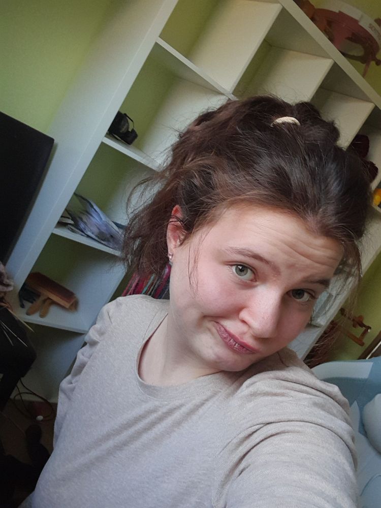 Ich vor einem weißen Regal vor einer grünen Wand. Ich habe ein altrosa Oberteil an und trage meine Haare in einem unordentlichen Zopf. Ich verziehe den Mund