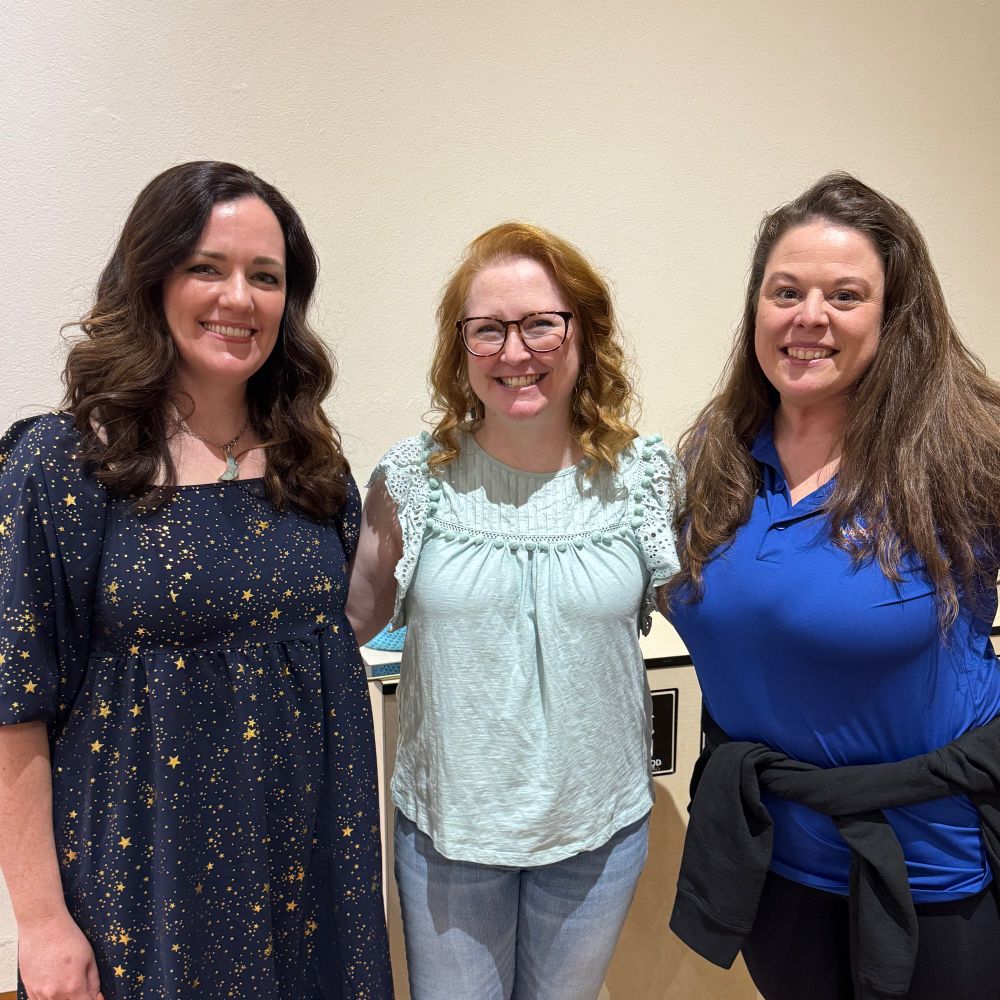 Three white women pose for the camera, smiling. Lindsay Lackey is on the left; she has long brown hair and wears a dark dress covered in stars. I’m in the middle; I have red hair and glasses, and am wearing a light turquoise blouse and jeans. On the right is Dr. Natasha Schatzman, a NASA engineer; she has long brown hair and is wears blue polo shirt with a jacket tied around her waist.