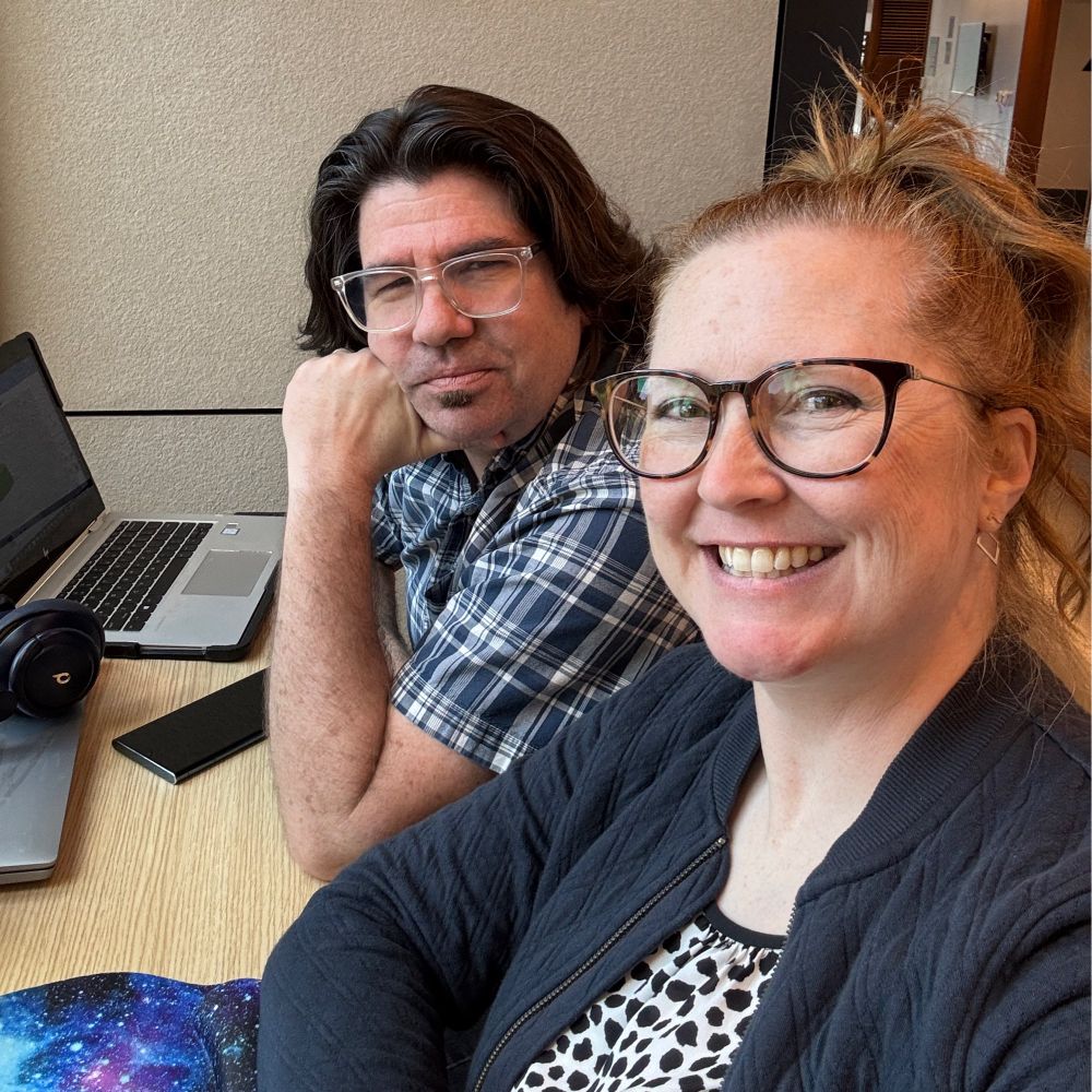 A white man and woman sit at a table with laptops out. The man has shoulder-length dark brown hair and glasses, and wears a blue plaid button-down shirt. The woman has curled red hair in a high ponytail, glasses, and a dark jacket open over a black and white spotted blouse.