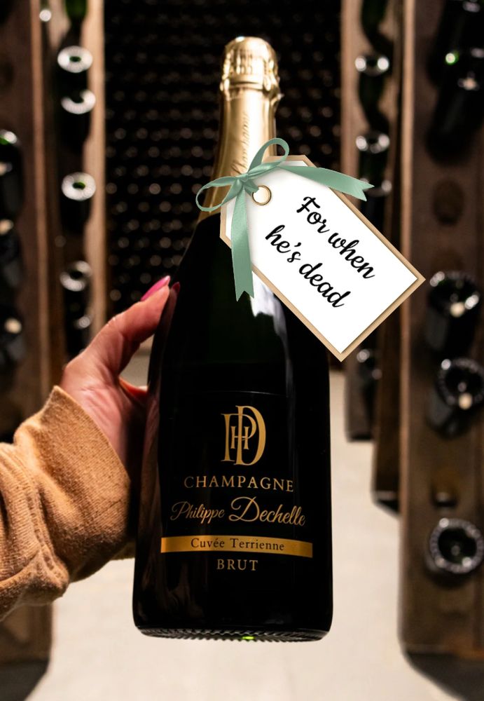 Image of a ladies hand holding a bottle of champagne with a gift tag that reads for when he’s dead. Background appears to be a wine cellar.