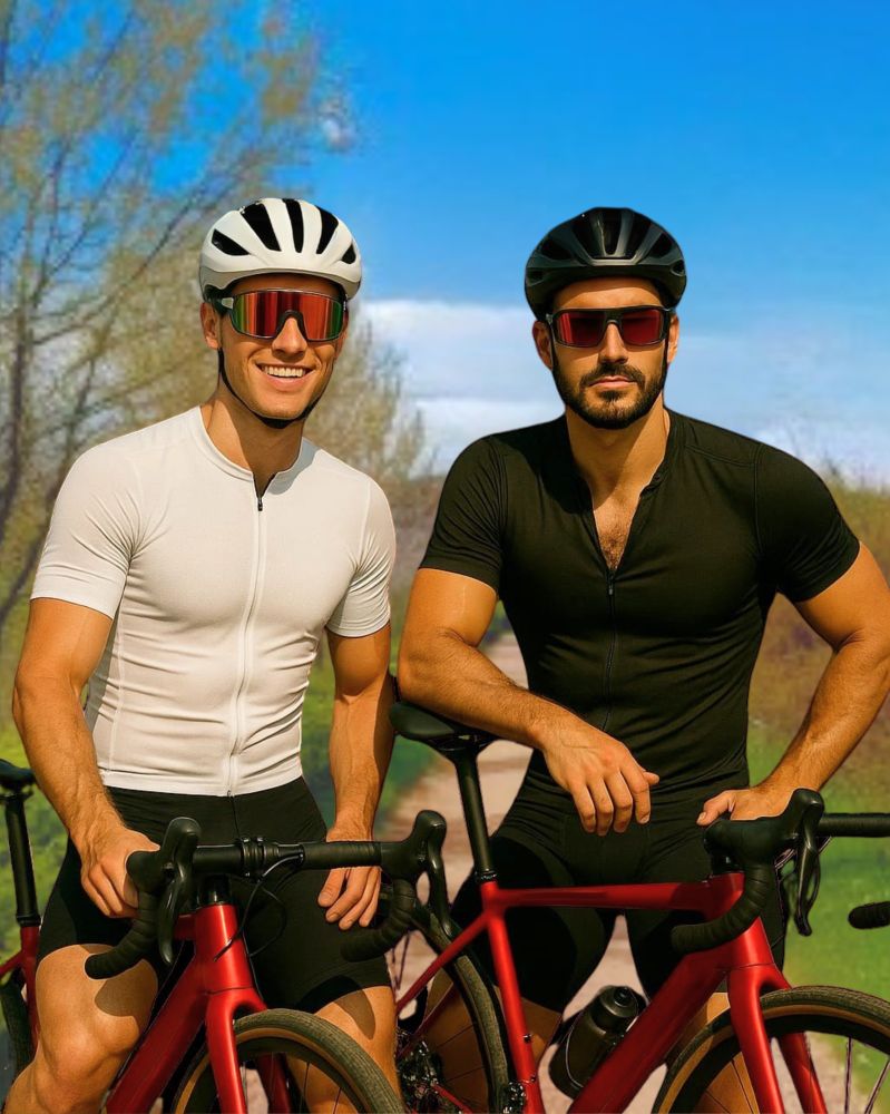 Zwei sportliche Männer in Radkleidung stehen nebeneinander auf ihren roten Gravelbikes. Beide tragen Fahrradhelme und reflektierende Sonnenbrillen. Der Mann links trägt ein weißes Trikot, der rechts ein schwarzes. Im Hintergrund ist ein klarer blauer Himmel mit einigen weißen Wolken zu sehen, dazu kahle Bäume und ein schmaler Schotterweg, der durch eine ruhige, ländliche Landschaft führt.