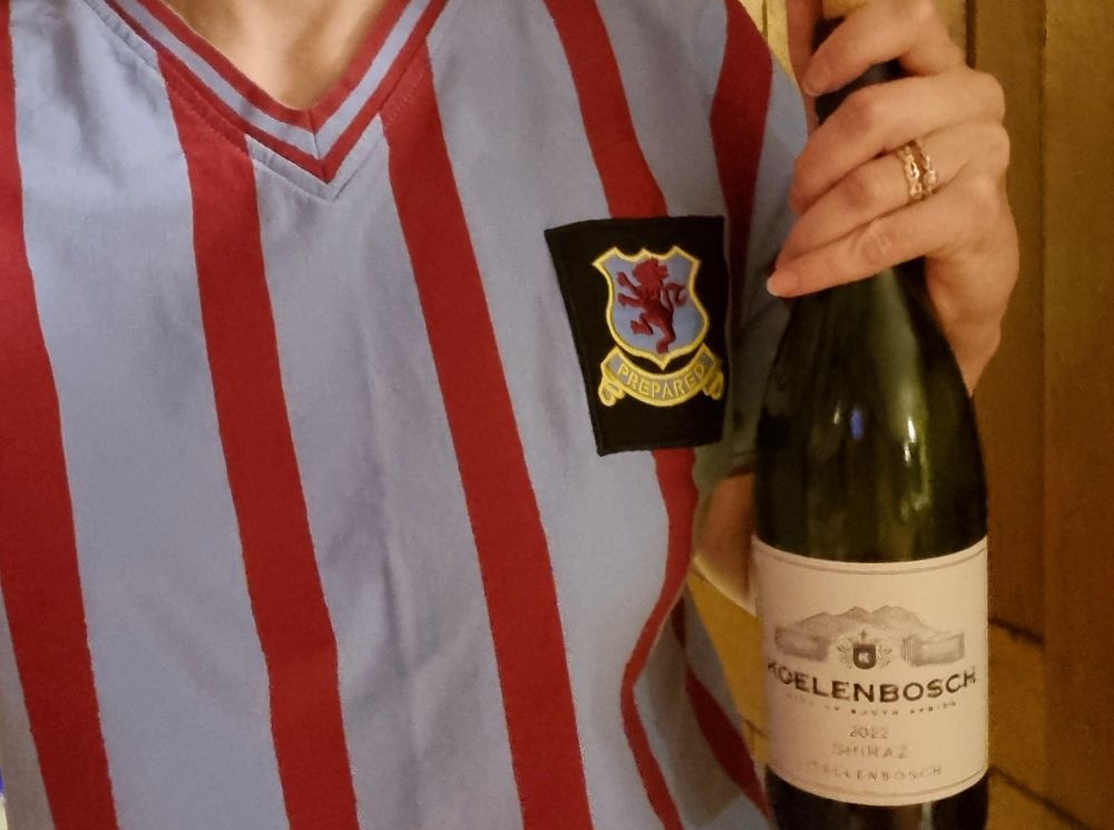 A hand holds a bottle of red wine (Koelenbosch Shiraz 2022) against a retro Aston Villa shirt with claret and blue stripes and a badge displaying a rampant lion above the motto 'Prepared'. 