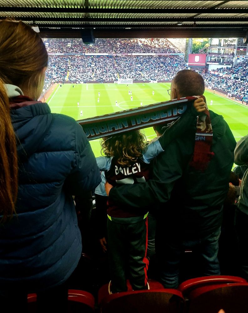 A child holds aloft an Aston Villa scarf with 'Morgan Rogers' embroidered on it whilst looking out over Villa Park during a match.